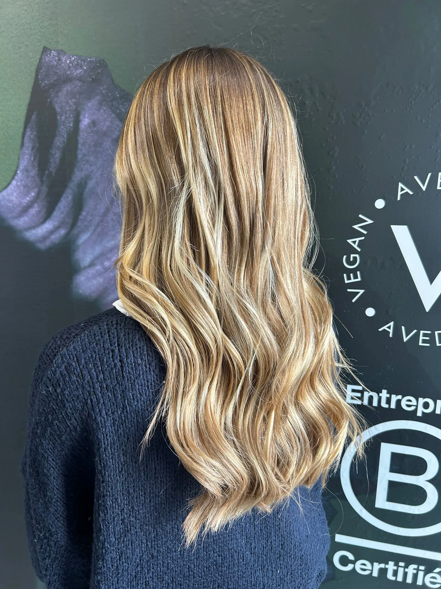 Chevelure blonde ondulée photographiée dans un salon de coiffure devant une signalétique Aveda
