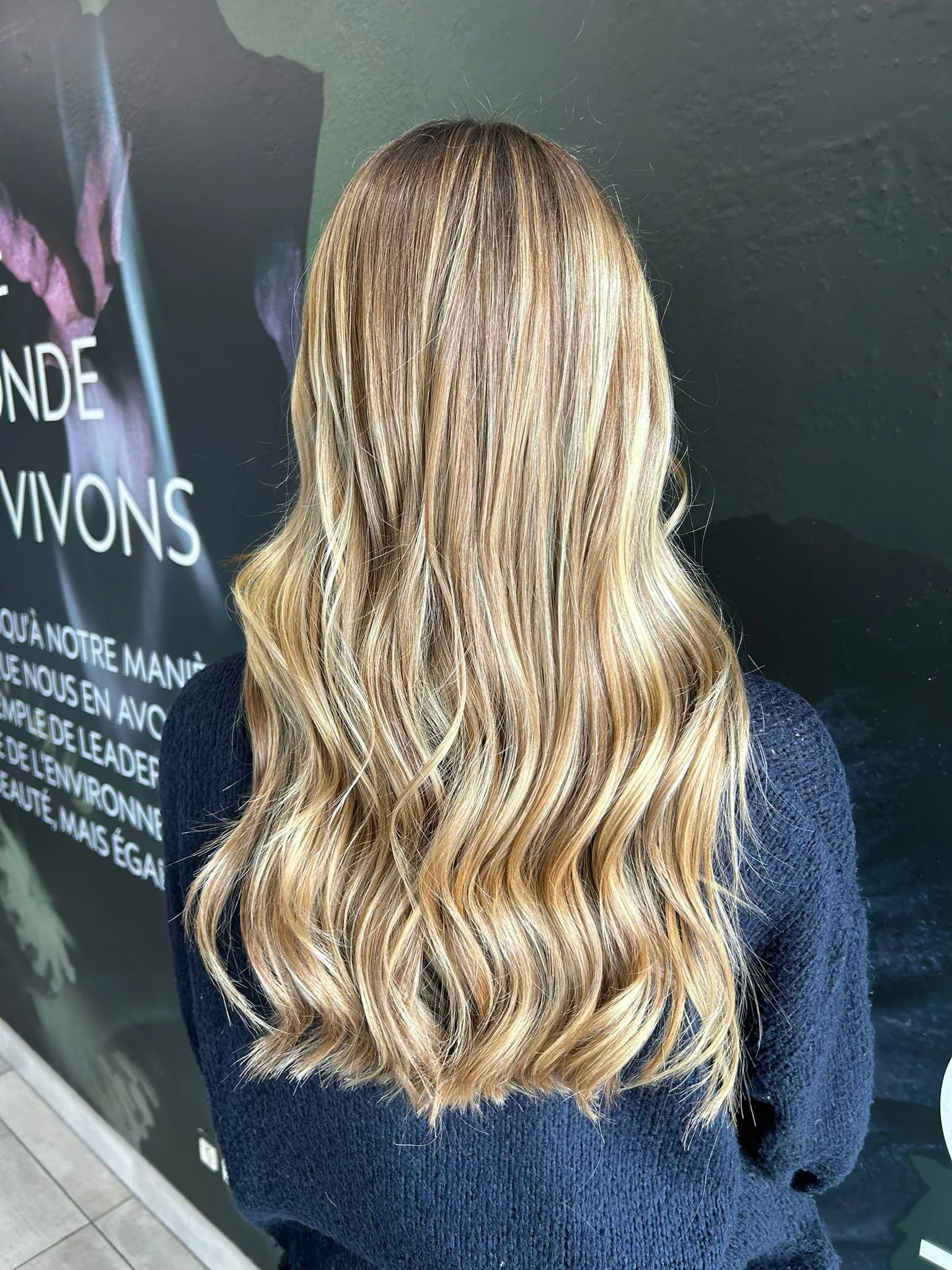 Vue de dos d’une chevelure longue et ondulée aux mèches blondes lumineuses, dans un salon à l’ambiance sombre
