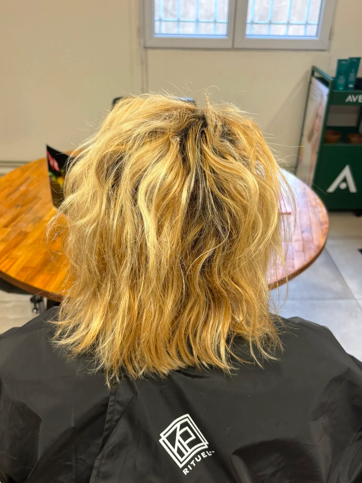 Chevelure blonde mi-longue et ondulée devant un présentoir Aveda